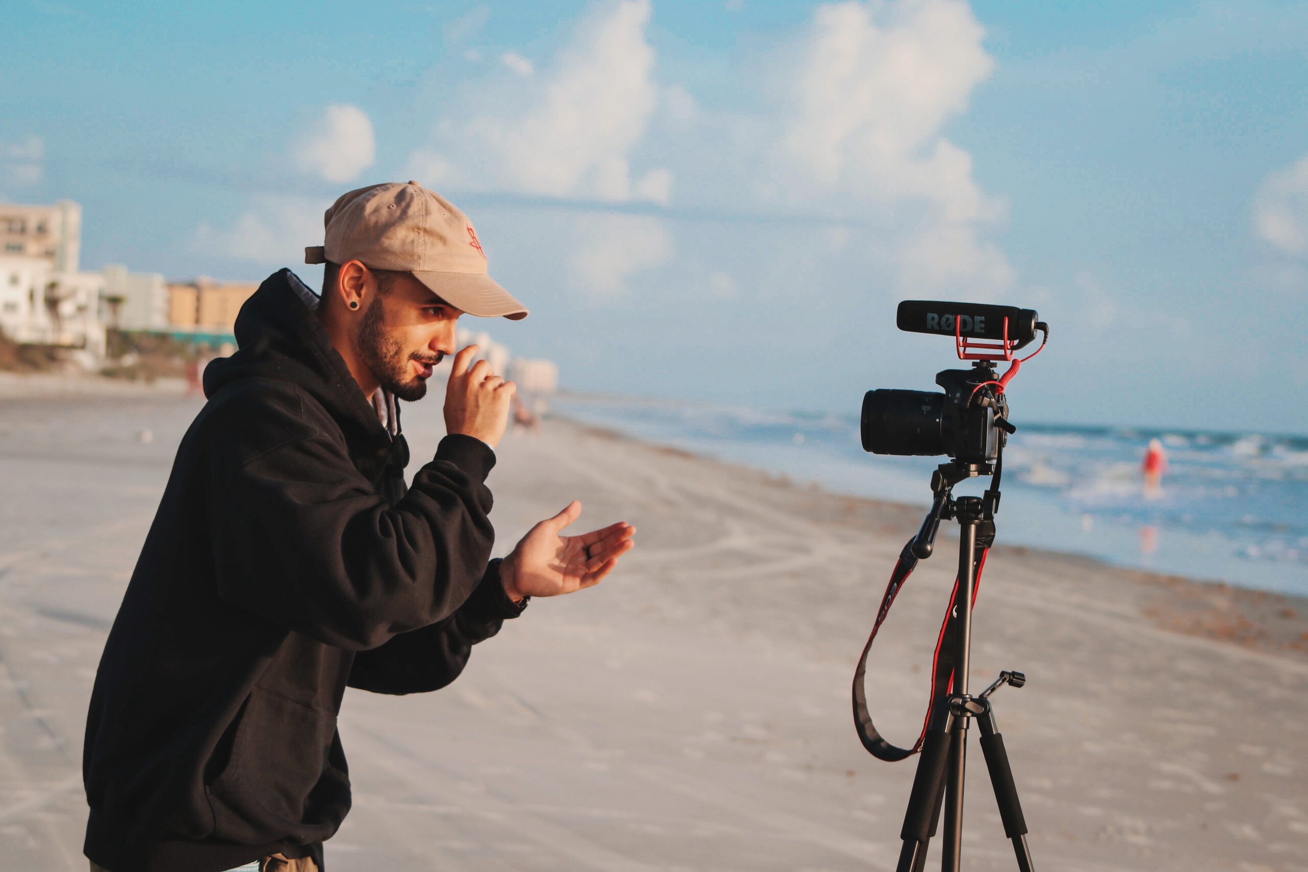 man in black jacket filming in front of camera on beach