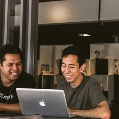 Two men sitting next to one another smiling and looking at a laptop