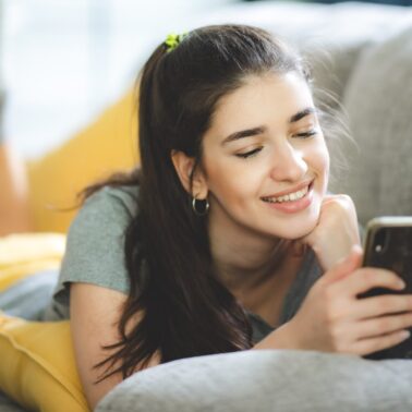 young woman laying on sofa and smiling while looking at phone